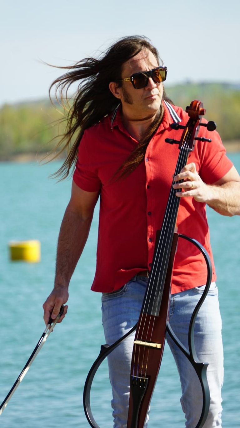 Mann in weißem Anzug spielt ein Cello in einer parkähnlichen Umgebung.