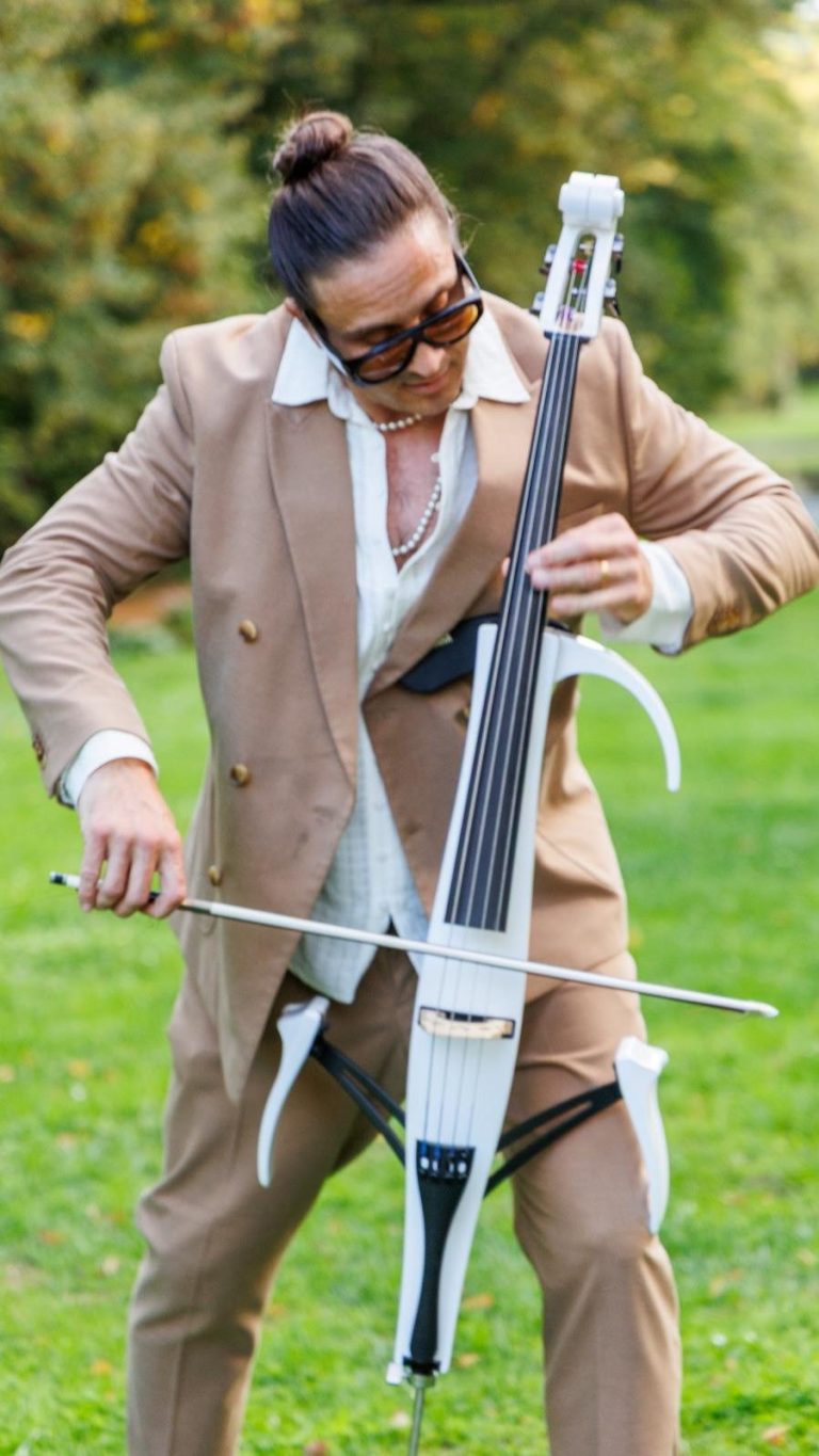Mann in weißem Anzug spielt ein Cello in einer parkähnlichen Umgebung.
