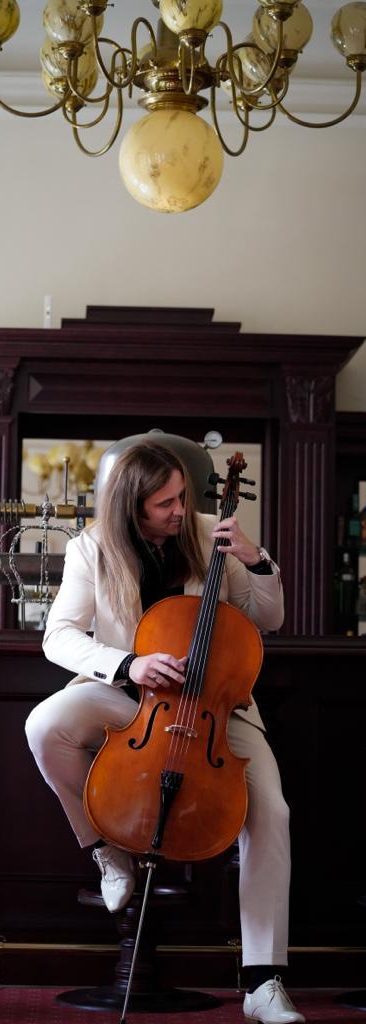 Ein Musiker in einem weißen Anzug spielt Cello in einem eleganten Raum.
